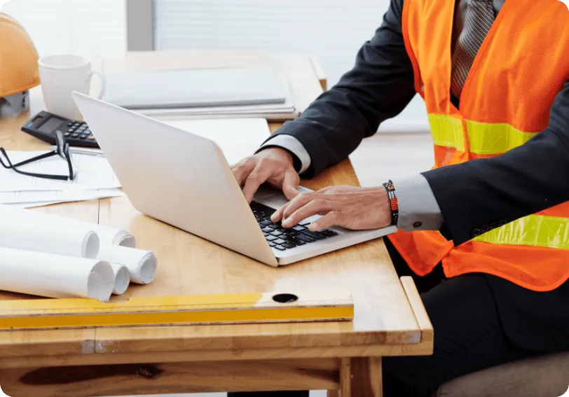 Person in a high-visibility vest working on a laptop next to building plans